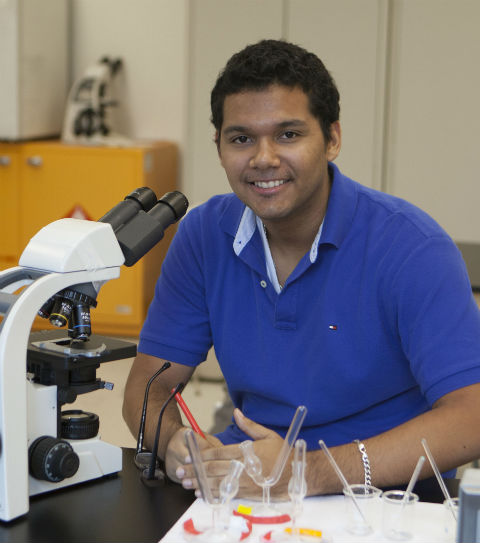 Teen sitting at a desk in front of a microscope smiling
